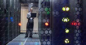 Technician with a laptop standing in a dim data center aisle between server racks with glowing status lights