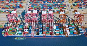 Aerial view of a busy container terminal with cargo ships, cranes and stacks of shipping containers.