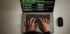 Hands typing on a laptop displaying green code, with a mouse beside it.