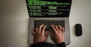 Hands typing on a laptop displaying green code, with a mouse beside it.
