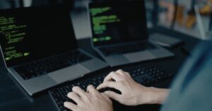 A person types on a keyboard in front of two laptops displaying code.
