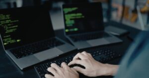 Hands typing on a laptop keyboard with lines of code on two laptop screens