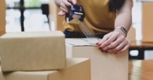 A person seals a cardboard shipping box with packing tape beside other parcels.