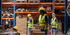 Shopify begins market-driven shipping rollout as rates move into Markets Warehouse workers in safety vests and hard hats handling boxes on storage racks, illustrating Shopify’s market-driven shipping rollout.
