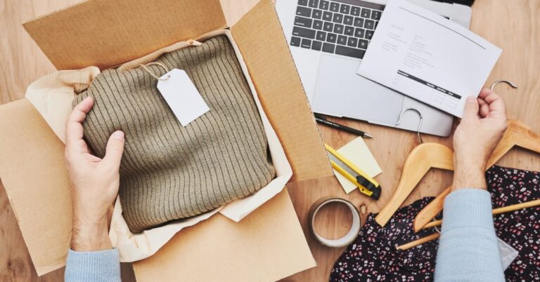 Business owner packing clothing in a shipping box beside a laptop and order paperwork.