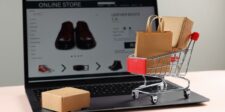 Shopify opens Shop MCP so AI agents can search, cart and check out Mini shopping cart with paper bags and a parcel on a laptop displaying an online store.